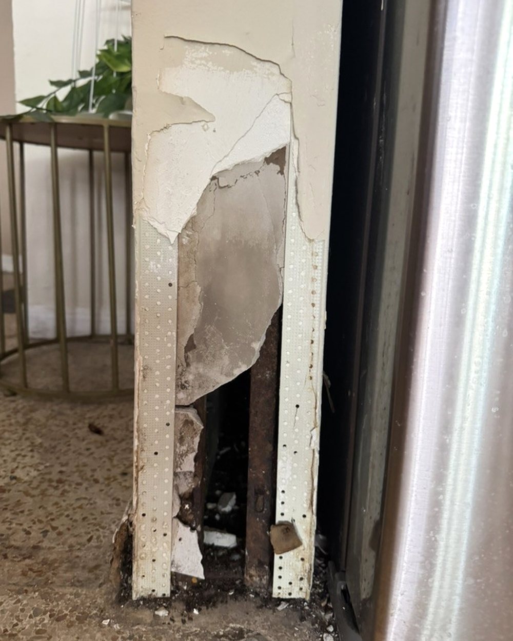 Damaged drywall and corner bead at the base of a wall beside a refrigerator
